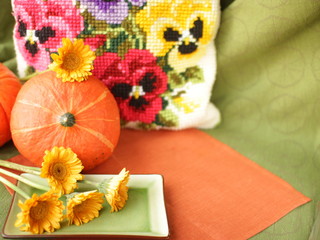 Orange pumpkins, yellow gerberas and a green rectangular plate on an orange linen napkin on a blurred background of pansies. Autumn still life on a green background. Place for text.