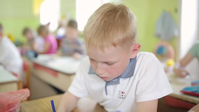 A red-haired boy with freckles draws a drawing on a white sheet while sitting at a table in kindergarten. 6 year old red-haired child is engaged in preschool education