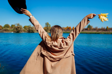 Autumn vibes. Young woman relaxing by river and having fun. Girl raising arms welcoming fall