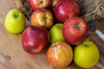 Red and green apples spilled out from a wicker basket lying on its side onto a brown wooden board, top side view, close-up