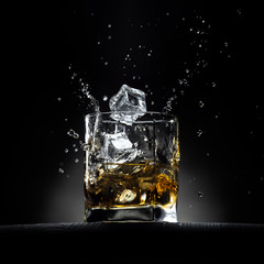 Glass of whiskey with ice cubes falling in it with splashes and drops. On the wooden table with dark background