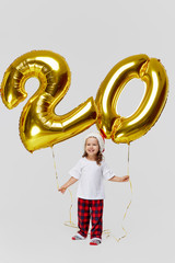 Portrait of little child girl in Santa hat posing with gold 20 twenty number balloons isolated over...