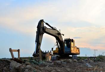 Heavy tracked excavator at a construction site on a background of a residential building and construction cranes on a sunny day against the backdrop of a sunset and blue sky with clouds