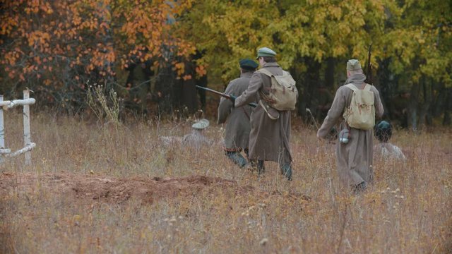 Several soldiers in warm coats walking on the field in front of the autumn forest - another soldiers sitting on the ground
