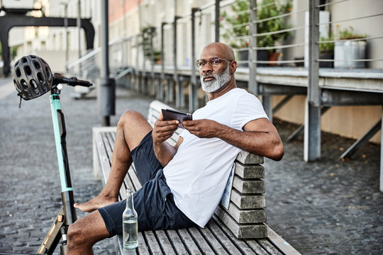 Portrait of mature, man with E-Scooter and cell phone sitting on bench in summer
