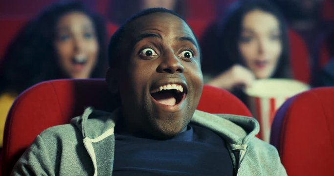 Close up of the cherful and happy African American young guy sitting in the cinema while watching nice action film and making surprised and satisfied face as loves this movie and looks enthusiastic.