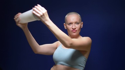 bald woman in a sports top shakes a cocktail in a shaker against a dark background.
