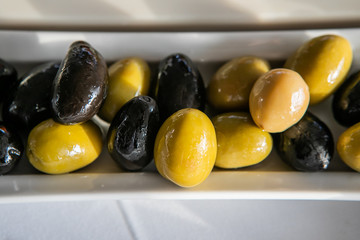 Variety Of Green And Black Marinated Olives Dish On Small Ceramic White Plate. Selective Focus. Close Up Of Tasty And Healthy Appetizer.