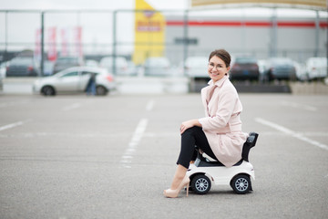 young woman with glasses in a supermarket parking lot sitting on a children's plastic car and...