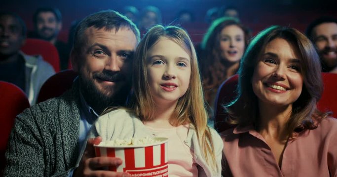 Portrait of the Caucasian happy mother and father sitting in the cinema with their pretty cute daughter while watching a film and eating popcorn.