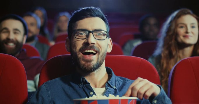 Close up of the cheerful young Caucasian attractive guy in glasses watching interesting film in the cinema and chewing popcorn.