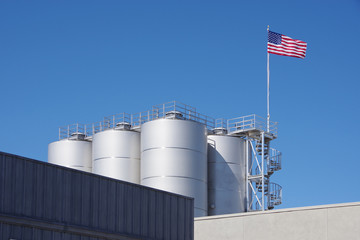 The US Flag waving above a group of large silos at an industrial facility under a bright blue summer sky