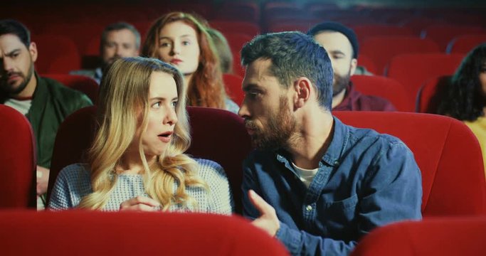 Young Caucasian pretty girl and handsome man sitting together at the date in the cinema and fighting because of the popcorn, then couple behind them asking to be quiet.