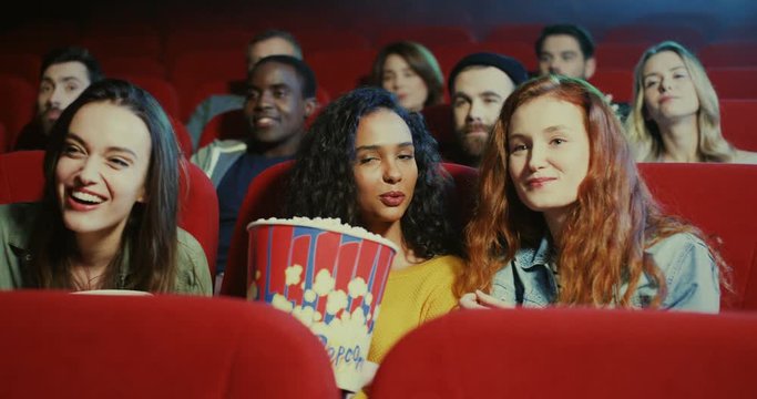 Three Caucasian cheerful and happy young beautiful girls eating popcorn, laughing and watching a comedy movie in the cinema.