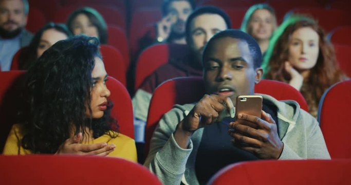 African American young handsome man tapping and scrolling on the smartphone while sitting in the cinema and his pretty moulatto girlfriend with popcorn in hands telling him to stop and watch a movie.
