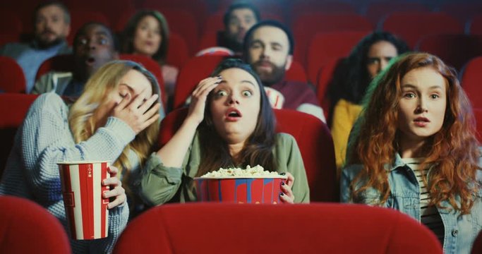 Three Caucasian beautiful girls best friends sitting with popcorn in the cinema and watching scary horror movie, then clothing eyes with hands as are afraid.