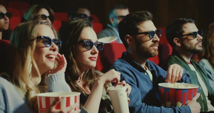 Two Caucasian young and beautiful women and two handsome men in 3D glasses with popcorn and drinks sitting in the cinema and watching an interesting action movie. Close up.