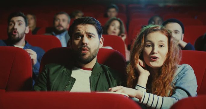 Close up of the young attractive Caucasian guy and girl watching a movie with popcorn together in the cinema, woman like it and man does not like.
