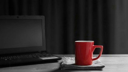 Focus on red coffee cup on napkins with blurred black laptop on wooden table and sunlight shine through the window in living room background,  coffee break concept, splash red color technique