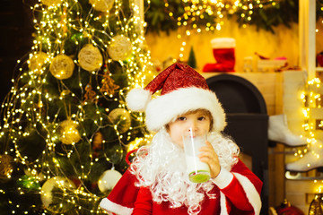 Fototapeta na wymiar Portrait of Santa Claus Drinking milk from glass and holding cookies. Little Santa Claus kid with beard and mustache. Santa boy in Santa hat.