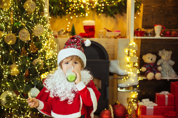 Kid Santa Claus enjoying in served gingerbread cake and milk. Greeting Christmas card. Happy Santa Claus - cute boy child eating a cookie and drinking glass of milk at home Christmas interior.