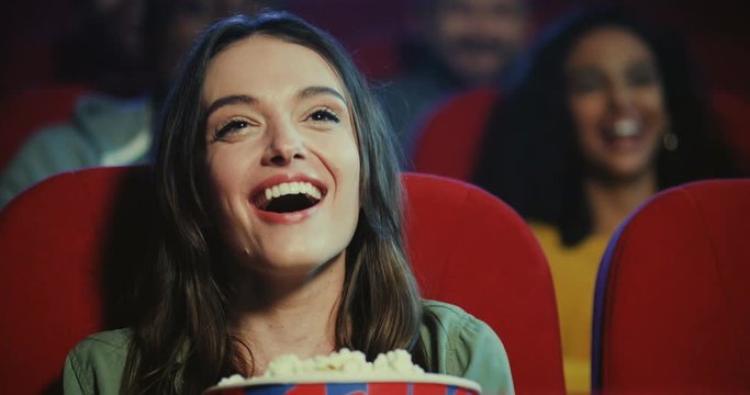 Portrait shot of teh funny young Caucasian attractive woman laughing aloud from a movie scene while watching a comedy film while sitting in the cinema with popcorn.