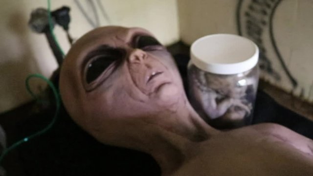Shot panning down at the alien laying on table with creature in jar next to him and green tubes connecting to the alien.