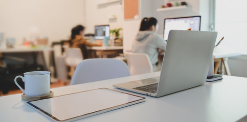 Laptop computer and tablet on white wooden table with office supplies with businesswomen in the background