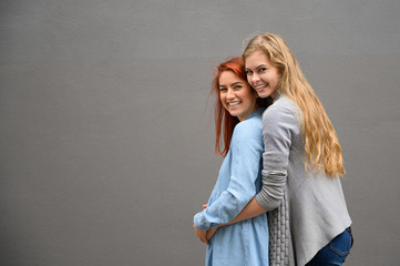 Same-sex relationships. A red-haired pregnant woman in a denim dress is standing against a gray wall, her hand is resting under her tummy. Her wife gently hugged the expectant mother from behind.