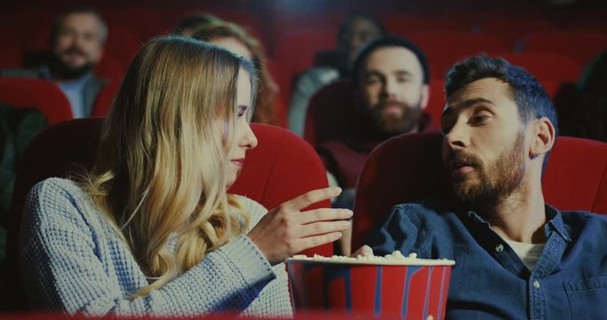 Young Caucasian blonde beautiful girl waking up her boyfriend while they watching a movie in the cinema and he telling the film is too boring so she feeding him with popcorn.