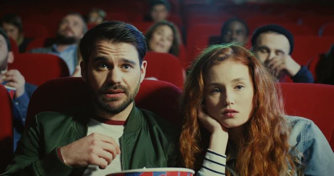 Close up of the young Caucasian man and womansitting in the cinema together at the date, eating popcorn and watching boring movie.