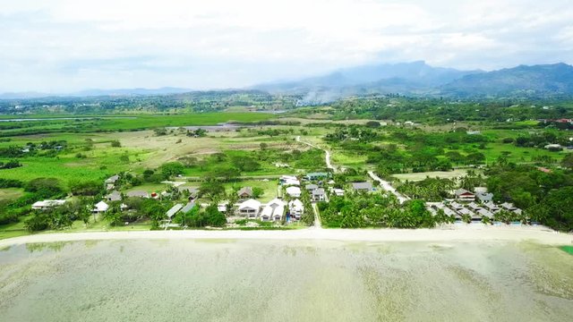 Aerial view: Sandy beachfront on pacific island with small houses, bures. Smoke from sugar cane fire and mountain in background