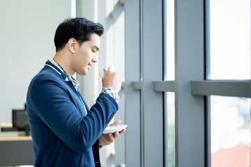 Portrait of smart and handsome young businessman drinking a coffee and looking outside of the window close up.