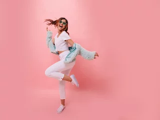 Fototapeten Inspiriertes positives Mädchen in weißen Turnschuhen, das auf rosafarbenem Hintergrund tanzt. Wunderschönes junges weibliches Modell mit dunklem, gewelltem Haar, das im Studio springt. Nicht isoliert. Platz kopieren. © ClaudiK