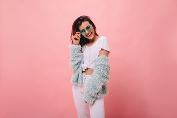 Excited white girl in bright stylish glasses posing on pink background. Dreamy curly woman playing with her ginger hair and laughing.