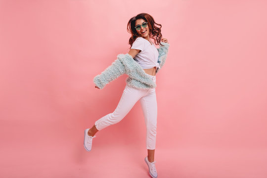  Inspired positive girl in white sneakers dancing on pink background. Gorgeous young female model with dark wavy hair jumping in studio. Not isolated. Copy space.