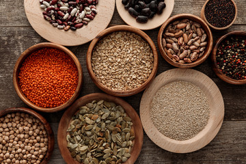 top view of bowls with raw oatmeal, red lentil, various beans, quinoa, chickpea, peppercorns and pumpkin seeds on wooden surface