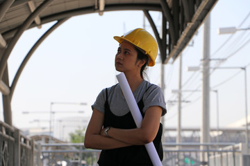 Female civil engineer or architect with yellow helmet, standing and hugging chest with project drafts while.
