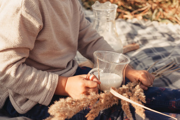 The child sits on a plaid and holds a glass of milk in his hands. A glass in the shape of an udder of a cow. Picnic in a wheat field. Milk advertisement for children