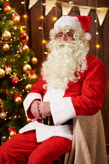 Santa Claus portrait, sitting indoor near decorated xmas tree with lights - Merry Christmas and Happy Holidays!