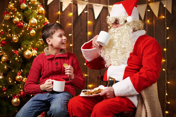 Santa Claus and child boy drinking tea, eating cookies, talking and sitting indoor near decorated xmas tree with lights - Merry Christmas and Happy Holidays!