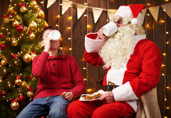 Santa Claus and child boy drinking tea, eating cookies, talking and sitting indoor near decorated xmas tree with lights - Merry Christmas and Happy Holidays!
