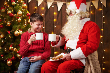 Santa Claus and child boy drinking tea, eating cookies, talking and sitting indoor near decorated xmas tree with lights - Merry Christmas and Happy Holidays!