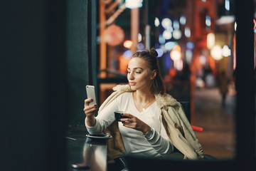 girl drinking coffee and using her phone outside a cafe during the evening