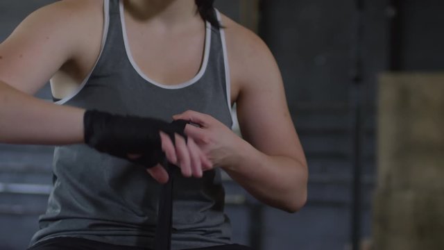 Mid-section shot of muscular female fighter sitting and wrapping hands for boxing in gym