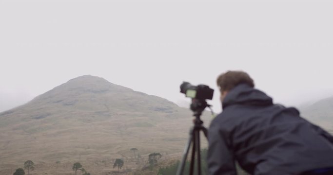 4K Camera Man in the Scottish Glen Coe Mountain Highlands. Tourist with tripod taking a photograph of hilly terrain and trees in Remote Scotland.