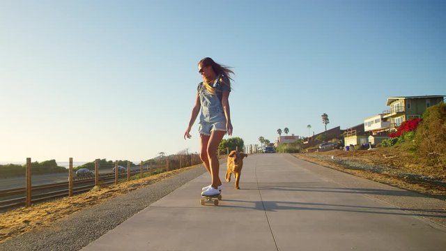 Young happy smiling girl skateboarding with golden retriever puppy at sunset 