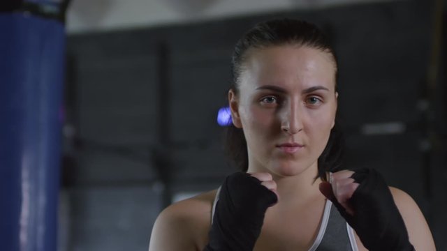 Portrait of professional female boxer with wrapped hands standing in defensive position and looking at camera in gym