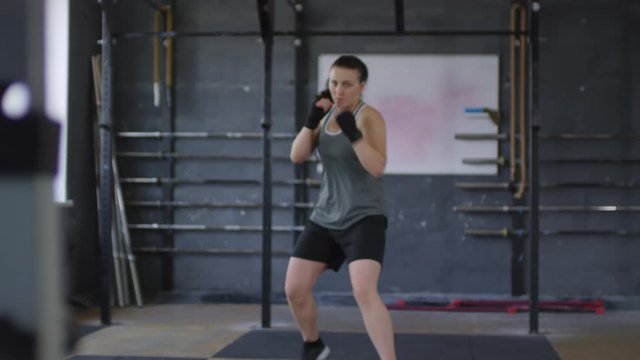 Rack focused shot of muscular female fighter with wrapped hands approaching camera and throwing punches in the air while having shadowboxing workout in gym