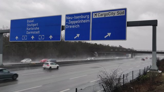 Establishment shot of moderate traffic moving during rainy days at the A5 Highway below the signage shows the direction to Cargocity S&uuml;d Airport in Frankfurt, Germany